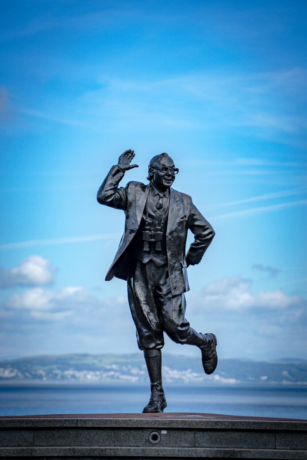 Bronze statue of a man in a jovial pose, standing on one leg with an arm raised, set against a clear blue sky backdrop. - Home Instead