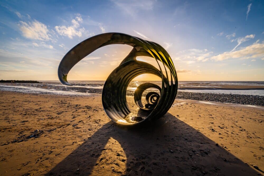 Sculpture of a spiral shell on a sandy beach during sunset with waves and cloudy sky in the background. - Home Instead