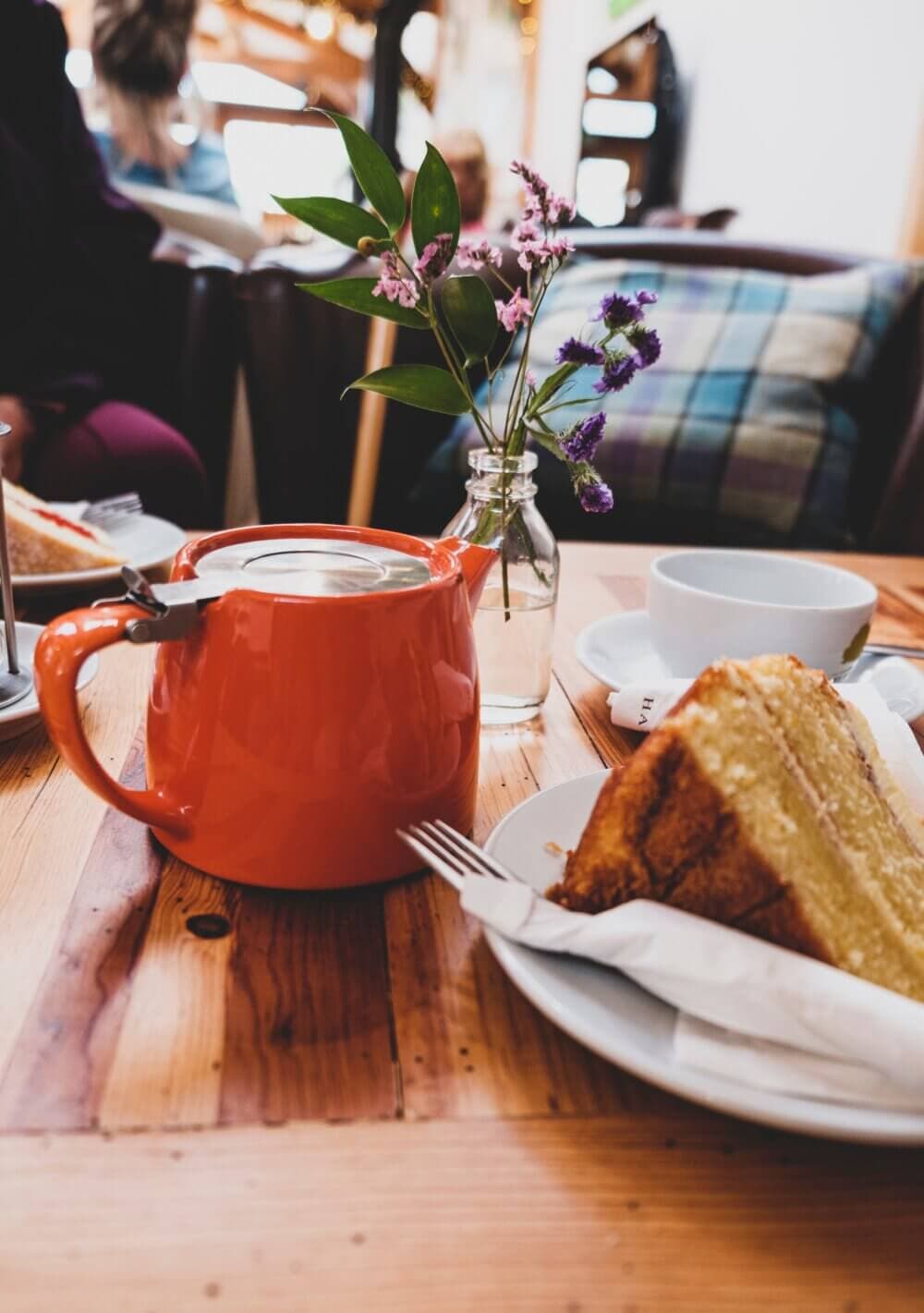 An orange teapot, a slice of cake, and a small vase with flowers on a wooden table in a cozy café setting. - Home Instead