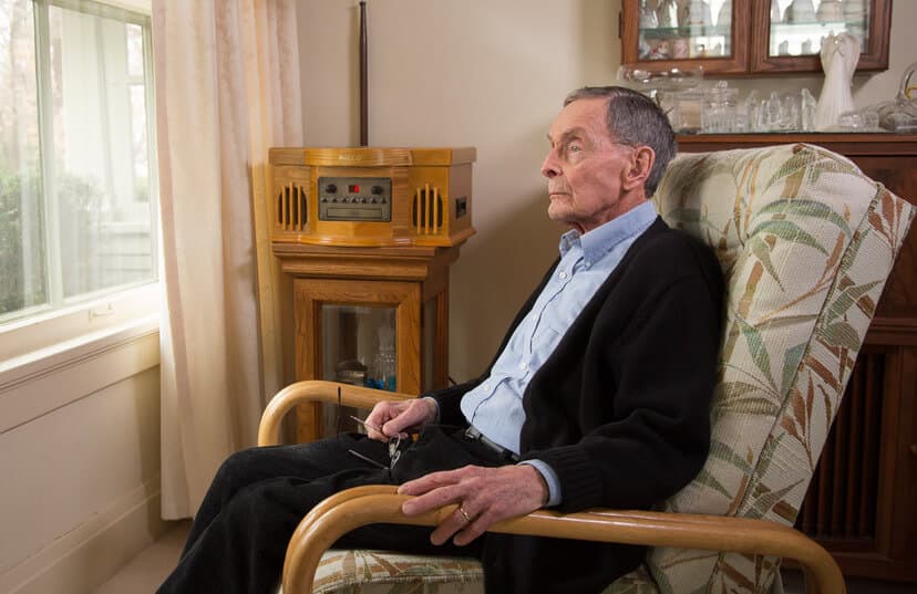 An elderly man in a blue shirt and black sweater sits in an armchair, holding glasses and looking out of the window. - Home Instead