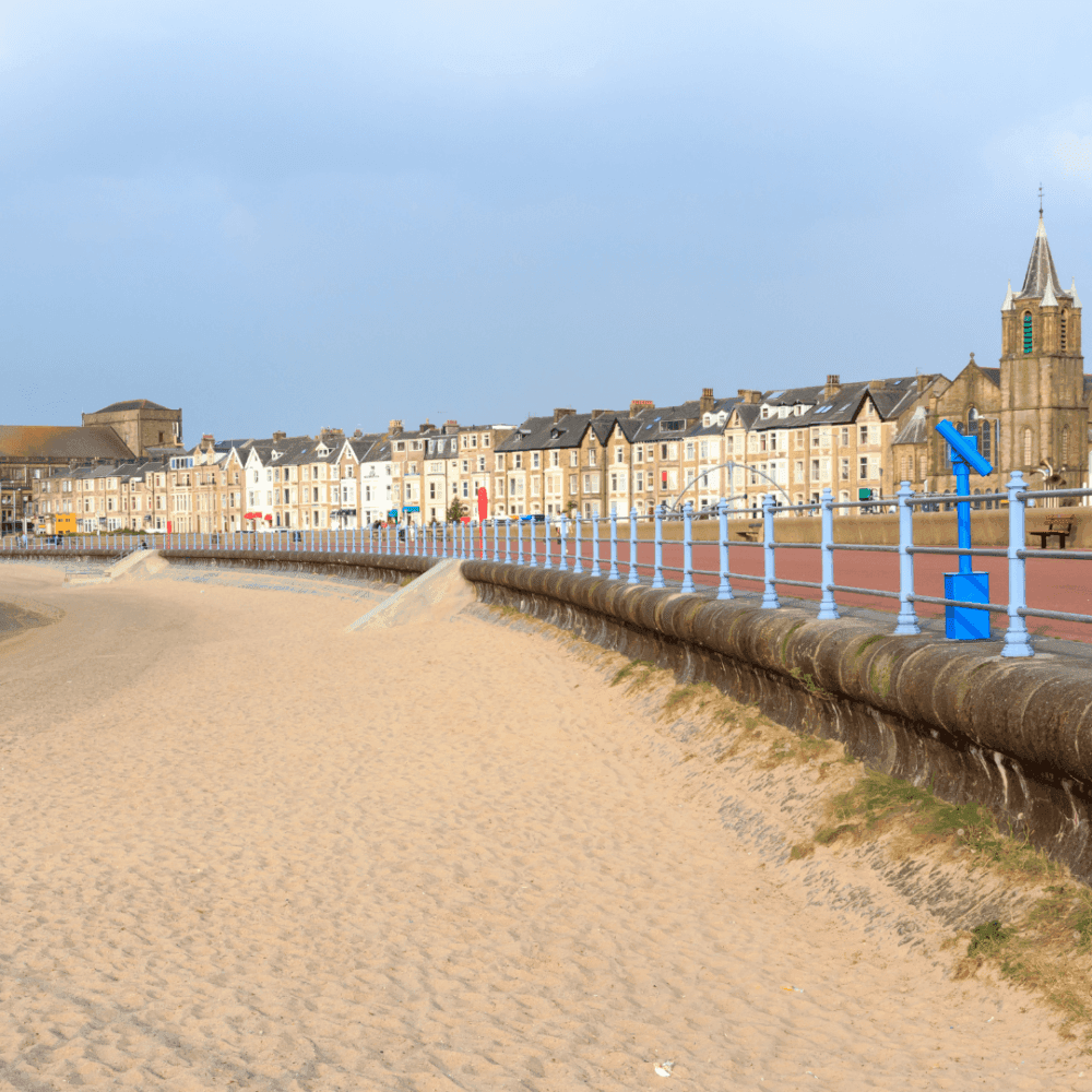 A sandy beach with a promenade lined with blue railings and a row of buildings in the background under a cloudy sky. - Home Instead