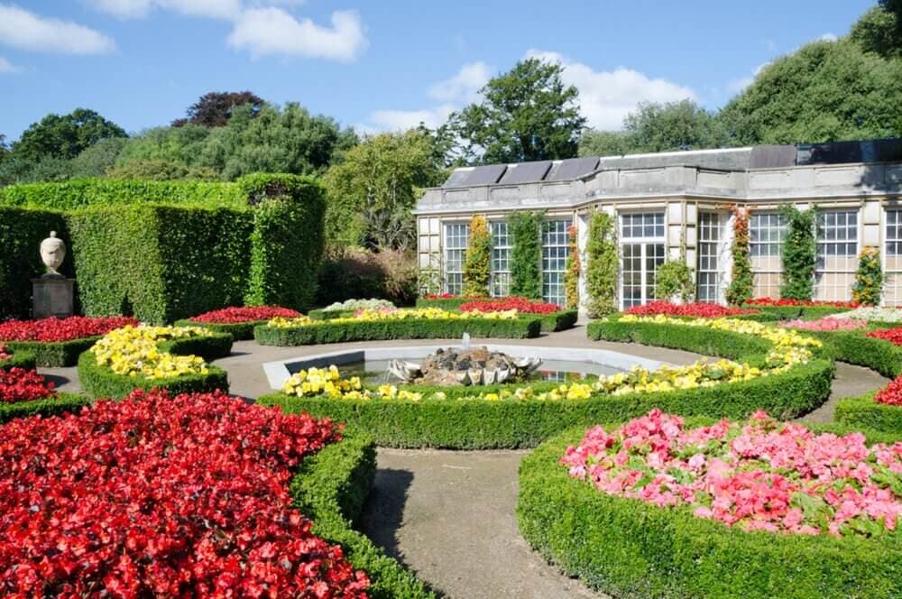 A formal garden with red, yellow, and pink flowers, neatly trimmed hedges, a central fountain, and a building in the background. - Home Instead