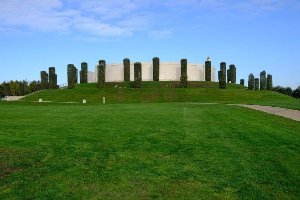 A circular memorial building on a hill, surrounded by tall, cylindrical trimmed trees, with a large grass field in front. - Home Instead