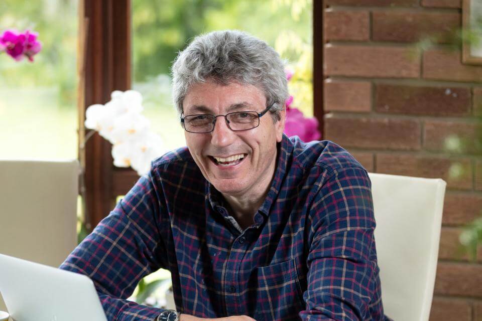 A smiling man with glasses, wearing a checkered shirt, sits at a table with a laptop in a bright room. - Home Instead