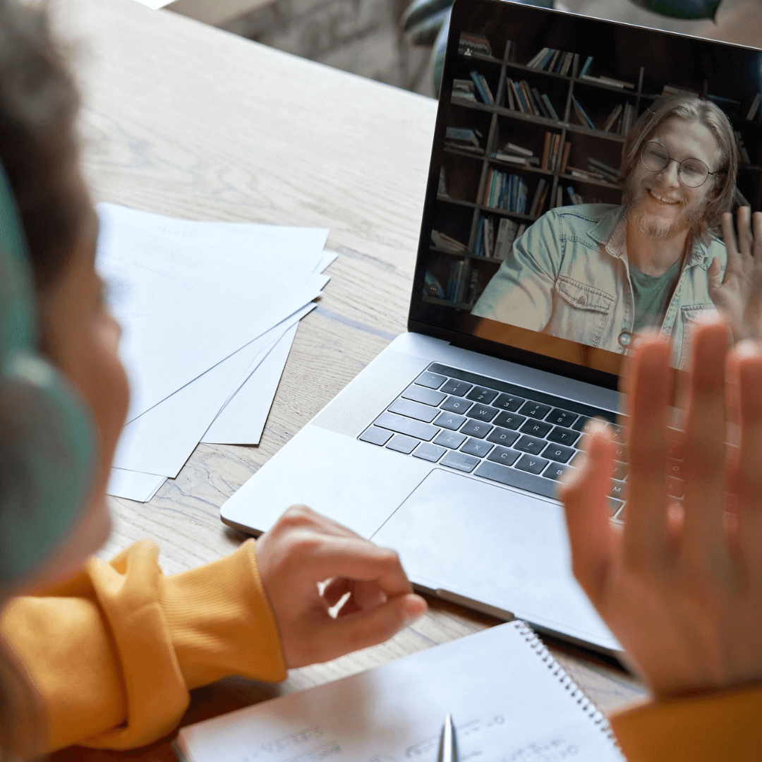 Person in headphones waves at a bearded man on a laptop screen during a video call, papers and notebook on desk. - Home Instead