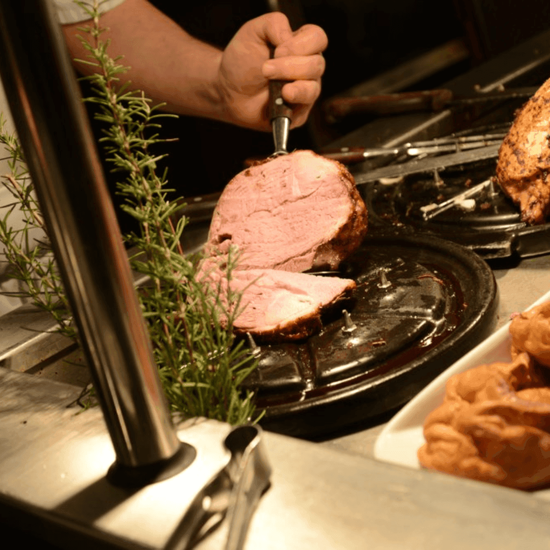Person slicing a piece of cooked meat on a black cutting board, with herbs and roasted bread in the foreground. - Home Instead