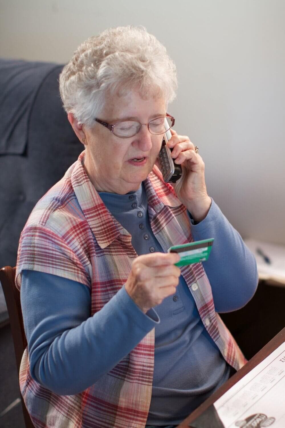 Elderly woman with glasses talking on a phone and holding a green credit card while sitting at a desk. - Home Instead