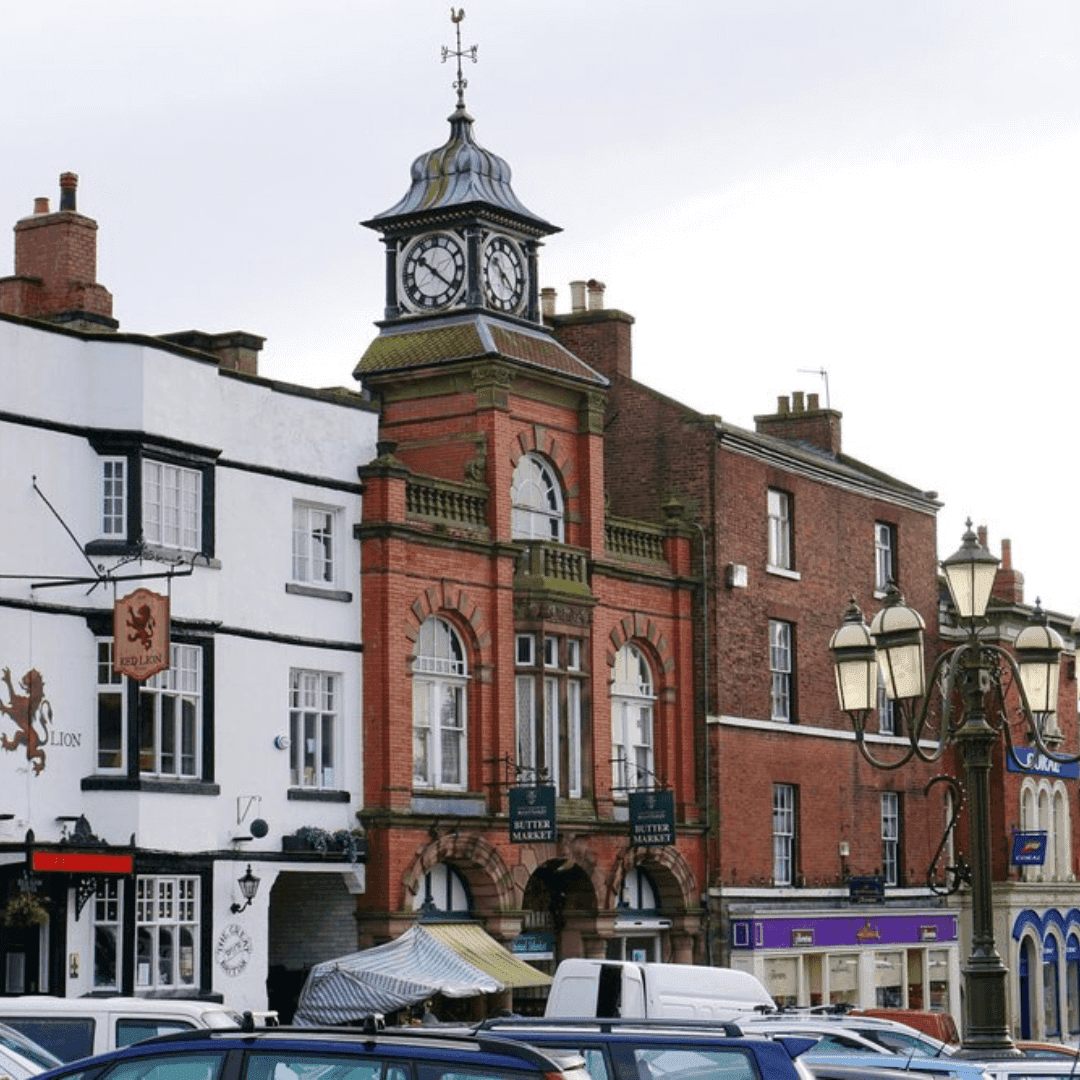 Town center with a prominent clock tower on a brick building, shops, parked cars, and a vintage lamppost in the foreground. - Home Instead