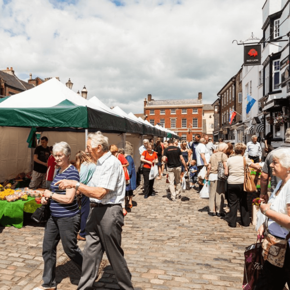 People walking and shopping at an outdoor market with stalls and a brick building in the background. - Home Instead