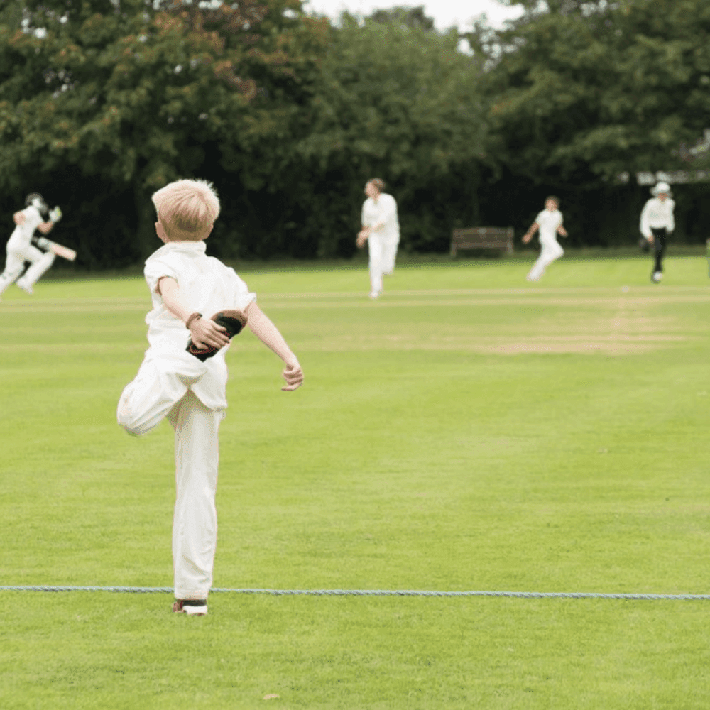 Child playing cricket, standing on one leg with one arm stretched back, facing other players on a field. - Home Instead