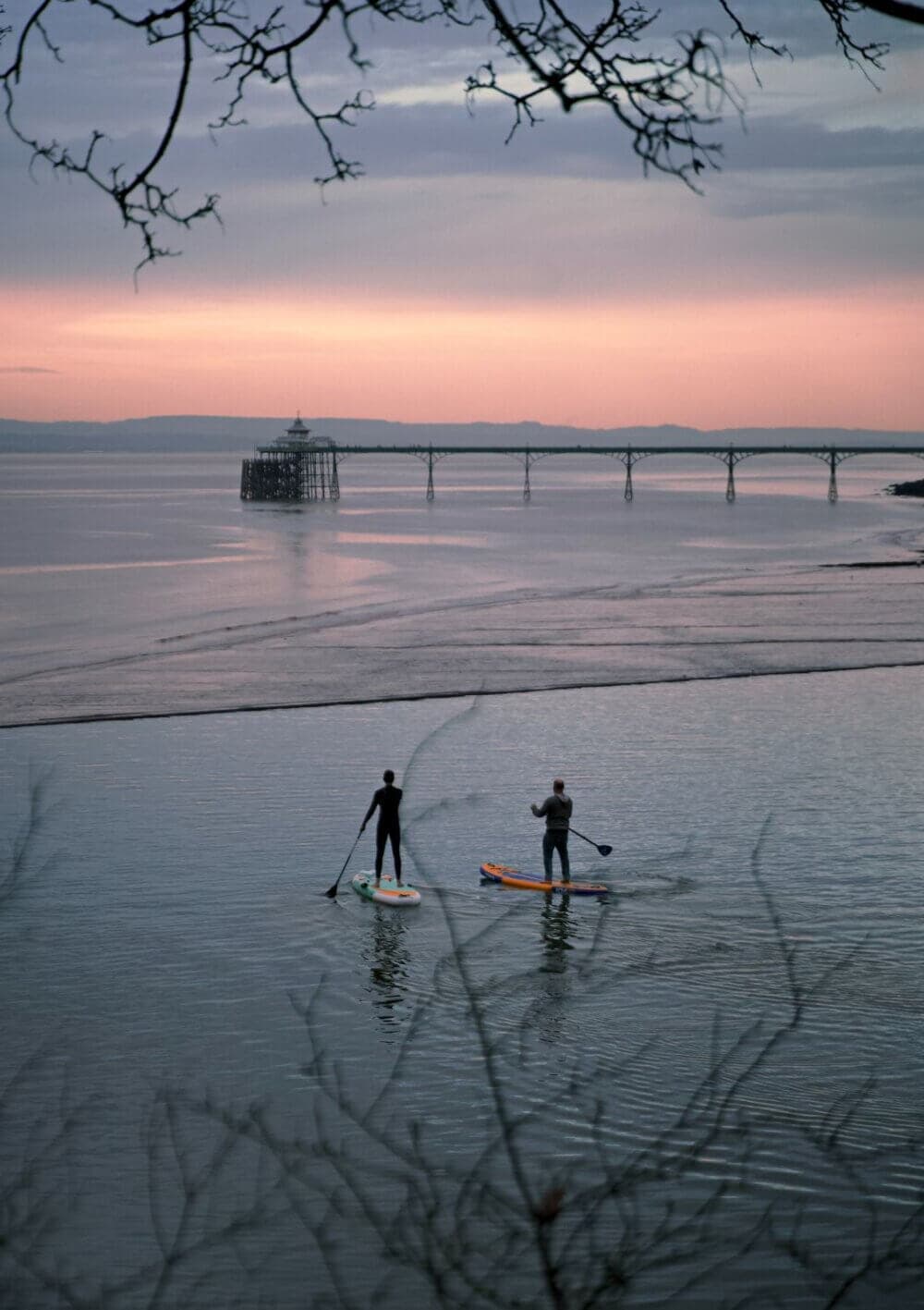 Two people paddleboarding on calm water at sunset, with a pier and distant mountains in the background. - Home Instead