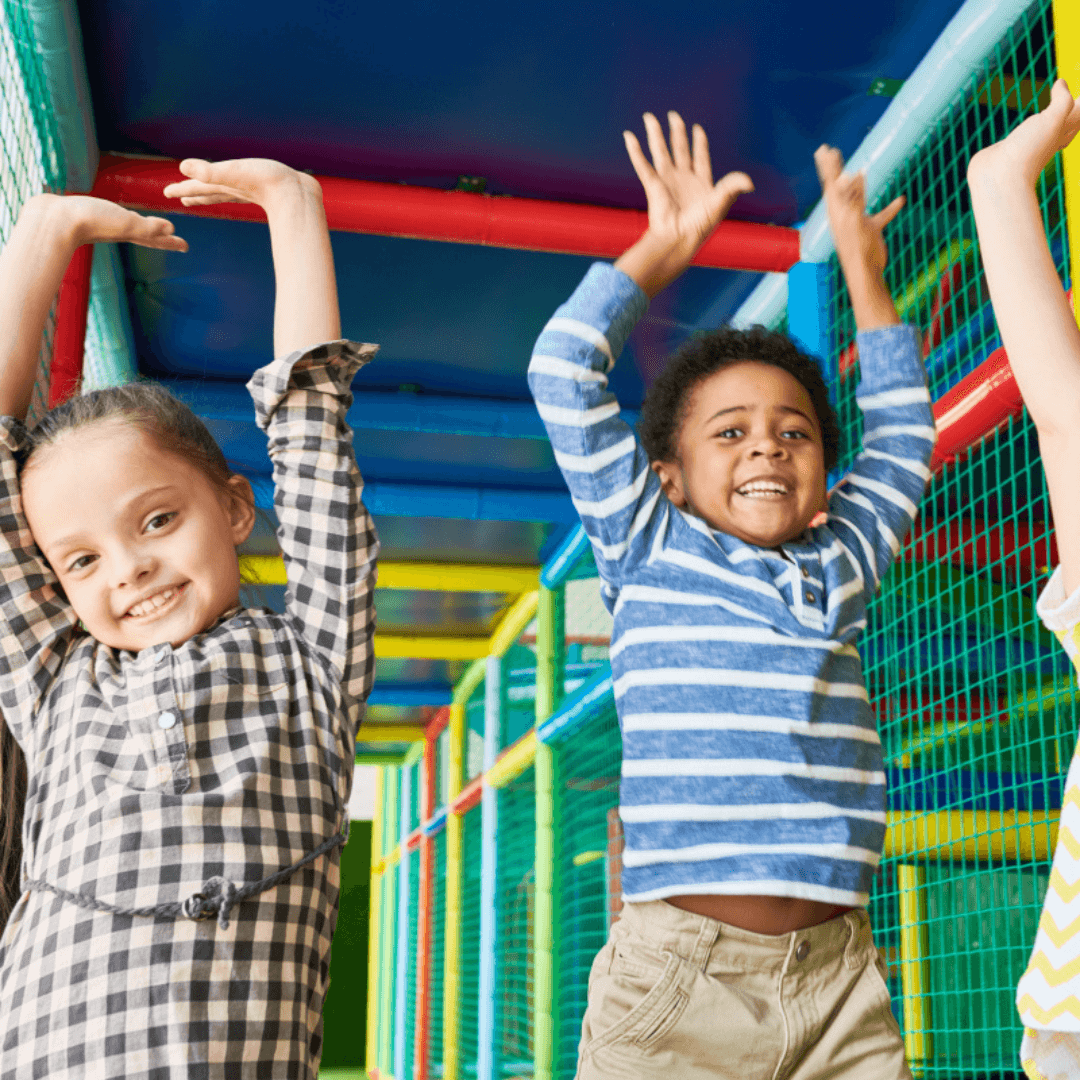 Three children raising their arms and smiling inside a colorful indoor play area with various tubes and nets. - Home Instead