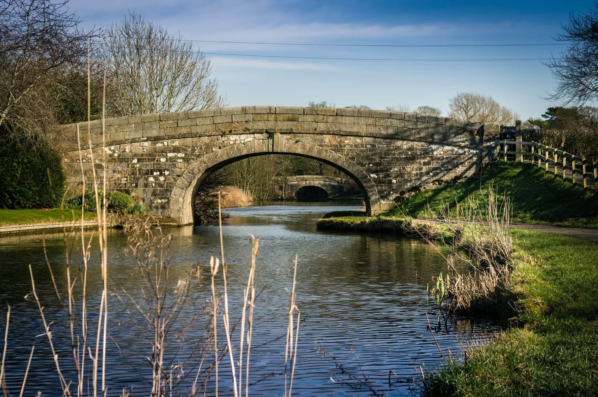 Stone bridge arching over a calm river, surrounded by trees and grassy paths under a clear blue sky. - Home Instead