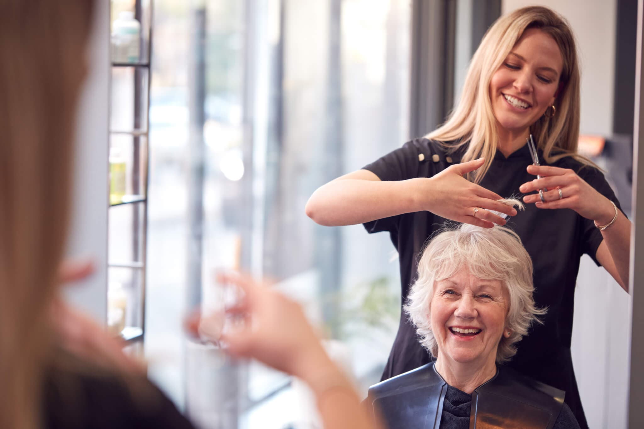 Hairdresser cutting an older woman's hair as they both smile in a salon, reflecting in the mirror. - Home Instead