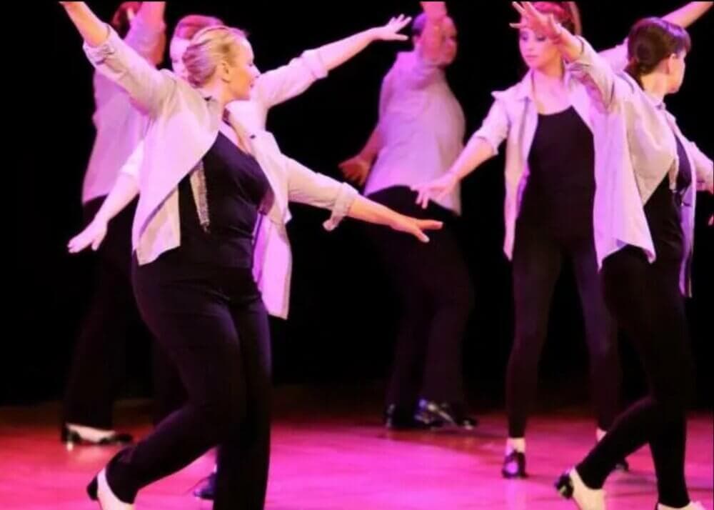 Dancers in white shirts and black pants performing on stage with outstretched arms and pink lighting. - Home Instead