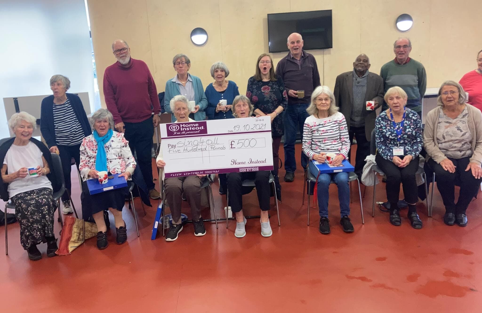A group of elderly people sitting, with some holding gifts and a large cheque for £500 from Home Instead in the center. - Home Instead