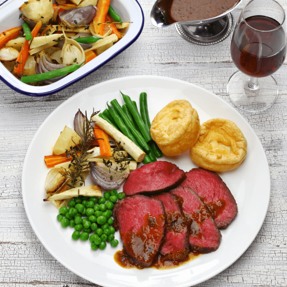 A plate with roast beef, Yorkshire puddings, green beans, peas, and gravy, with a side of roasted vegetables and a glass of wine. - Home Instead