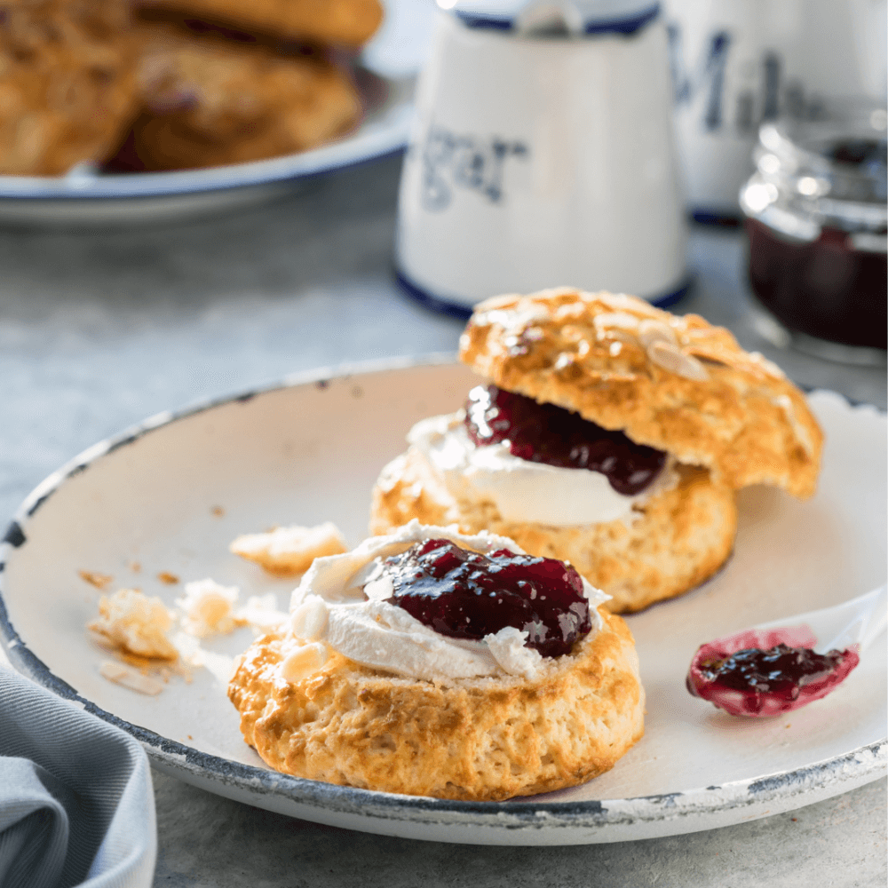A scone cut in half with cream and red jam on top, placed on a white plate with crumbs and a spoonful of jam beside it. - Home Instead