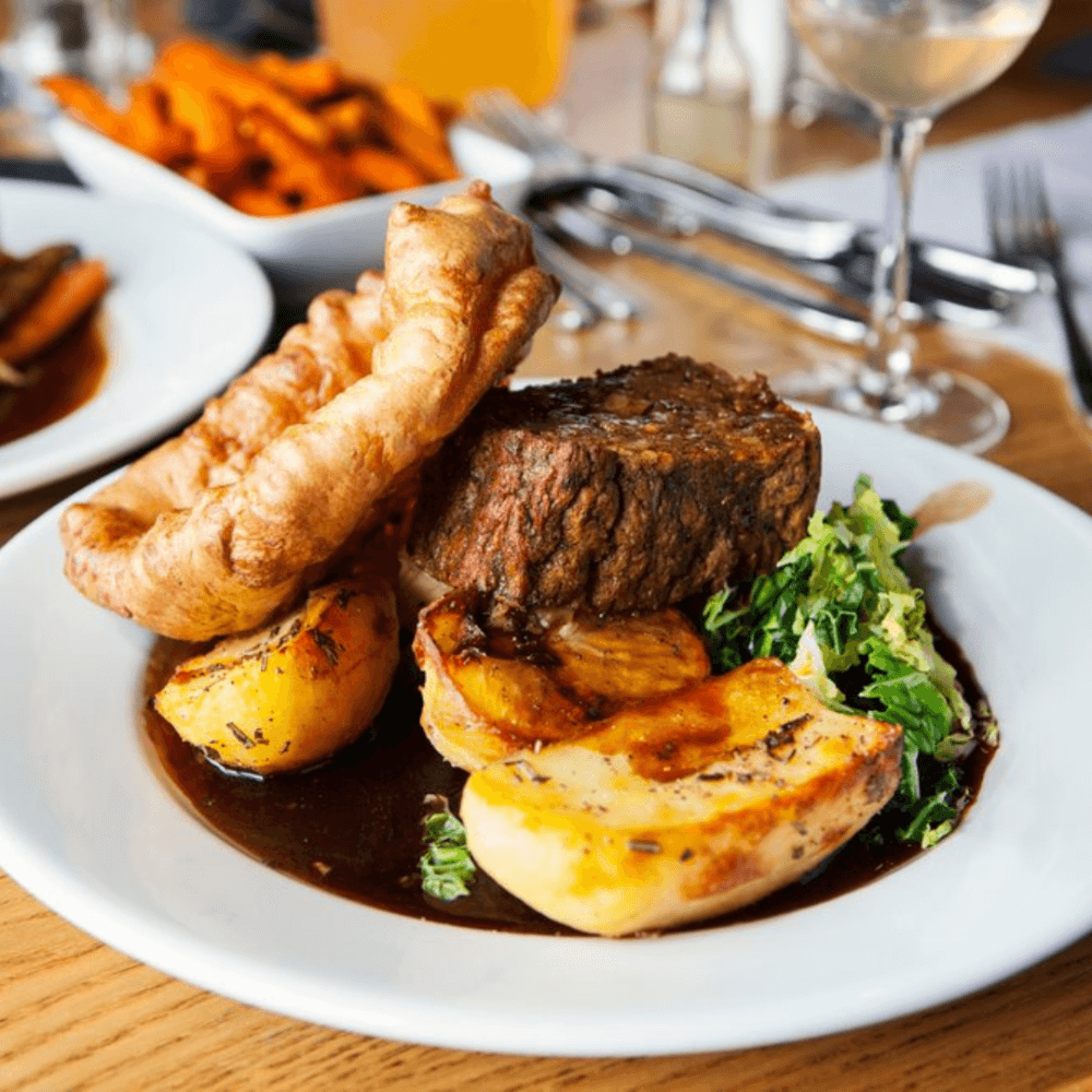 A plated meal featuring roast beef, Yorkshire pudding, roasted potatoes, and green vegetables with a glass of wine nearby. - Home Instead