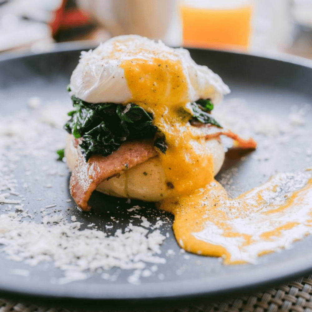 A plate with eggs benedict topped with spinach, bacon, and hollandaise sauce, with grated cheese sprinkled around. - Home Instead