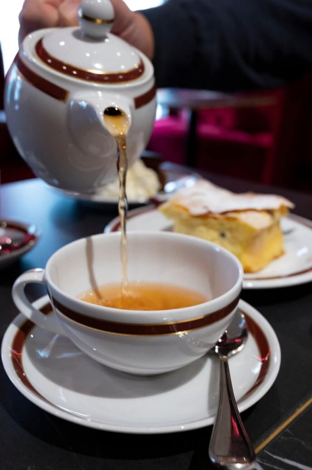 Teapot pouring tea into a cup on a saucer with a spoon, cake in the background. - Home Instead
