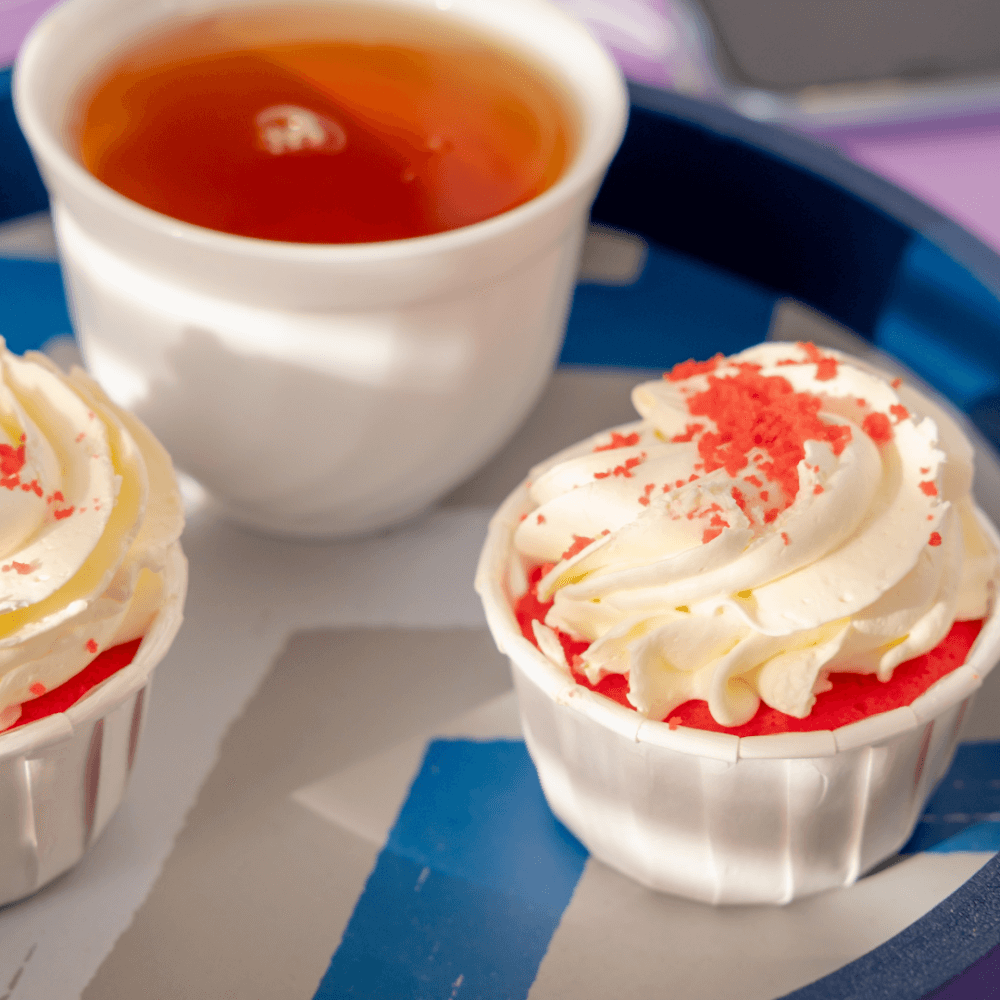 Two red velvet cupcakes with white frosting on a blue tray next to a cup of tea. - Home Instead