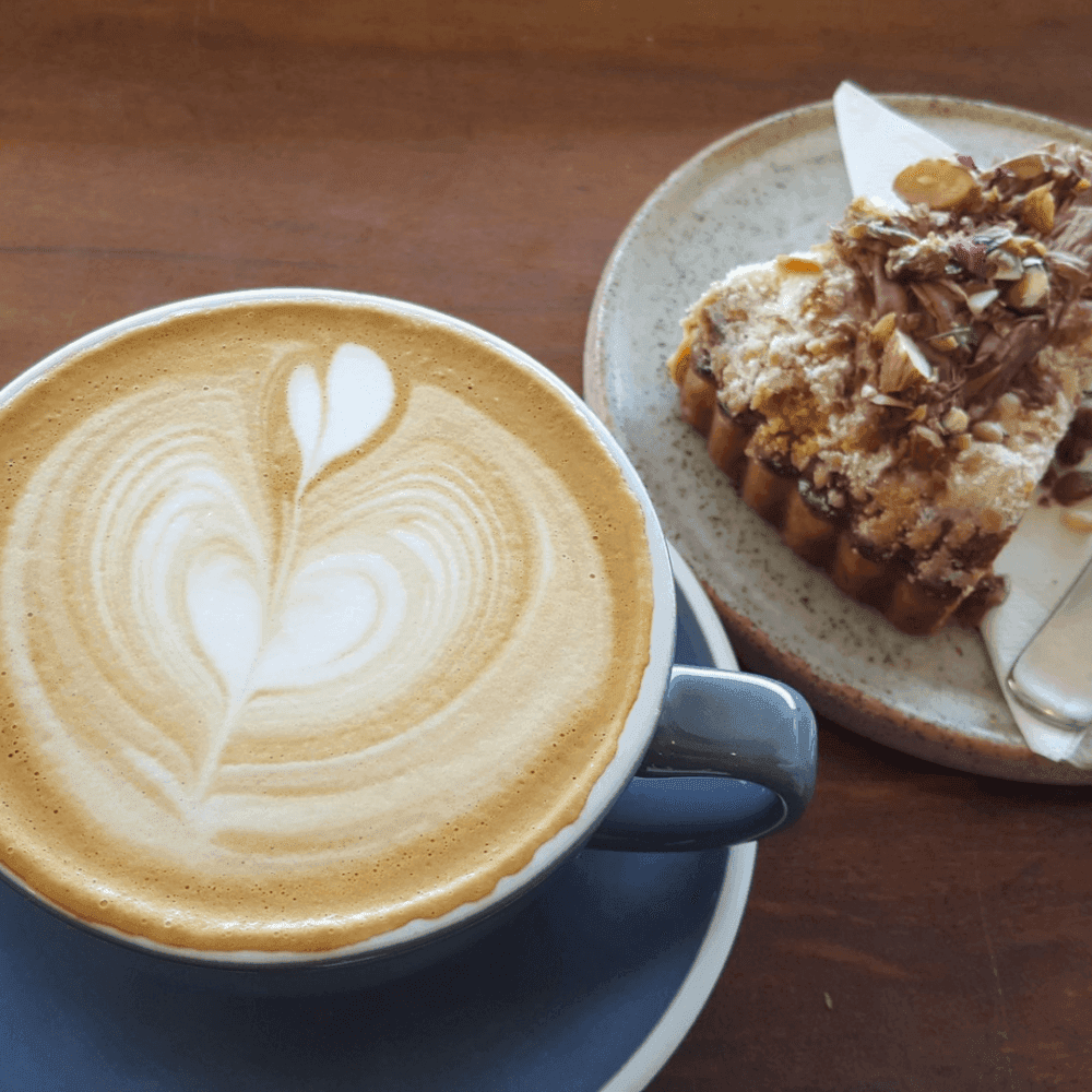 A latte with heart foam art in a gray cup and saucer next to a piece of dessert on a small plate with a napkin. - Home Instead