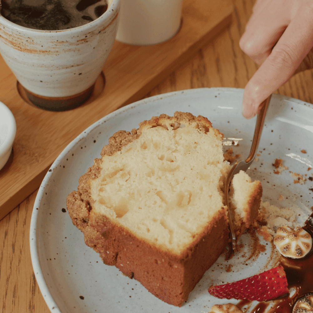 A hand cutting a slice of cake with a fork beside a cup of coffee and a plate with strawberries and chocolate drizzle. - Home Instead