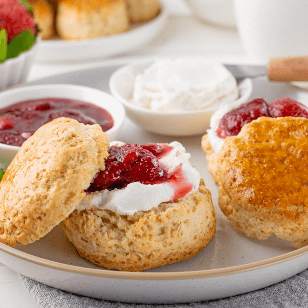 Scones with clotted cream and strawberry jam on a plate, accompanied by bowls of cream and jam in the background. - Home Instead