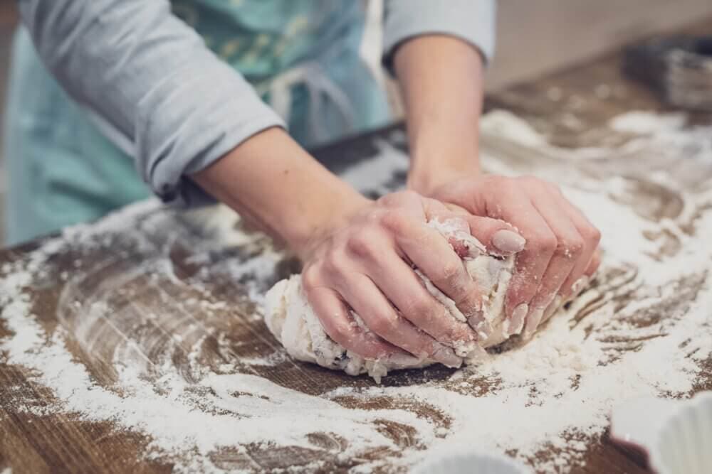 Hands kneading dough on a floured wooden surface in a kitchen setting. - Home Instead