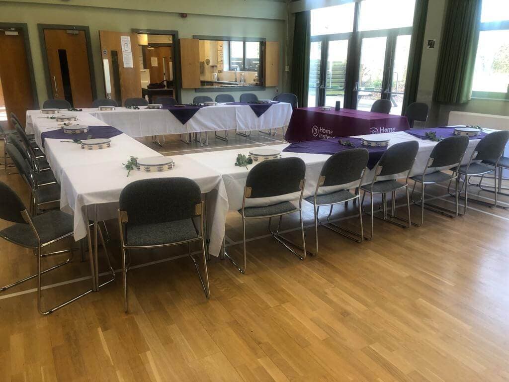 Large room arranged with tables covered in white and purple cloths, set with plates and chairs around them. - Home Instead