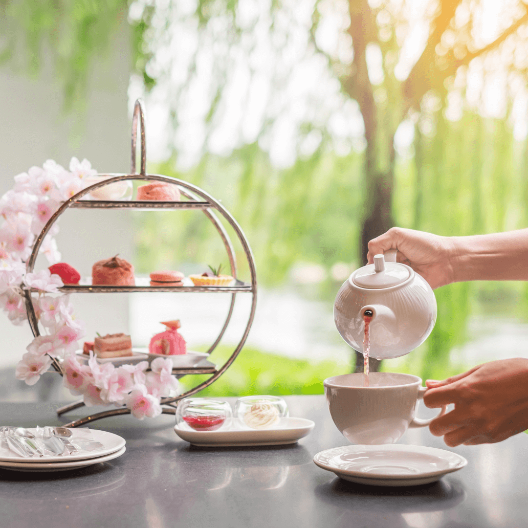 A person pours tea into a cup next to a tiered stand with assorted pastries and a scenic garden view in the background. - Home Instead
