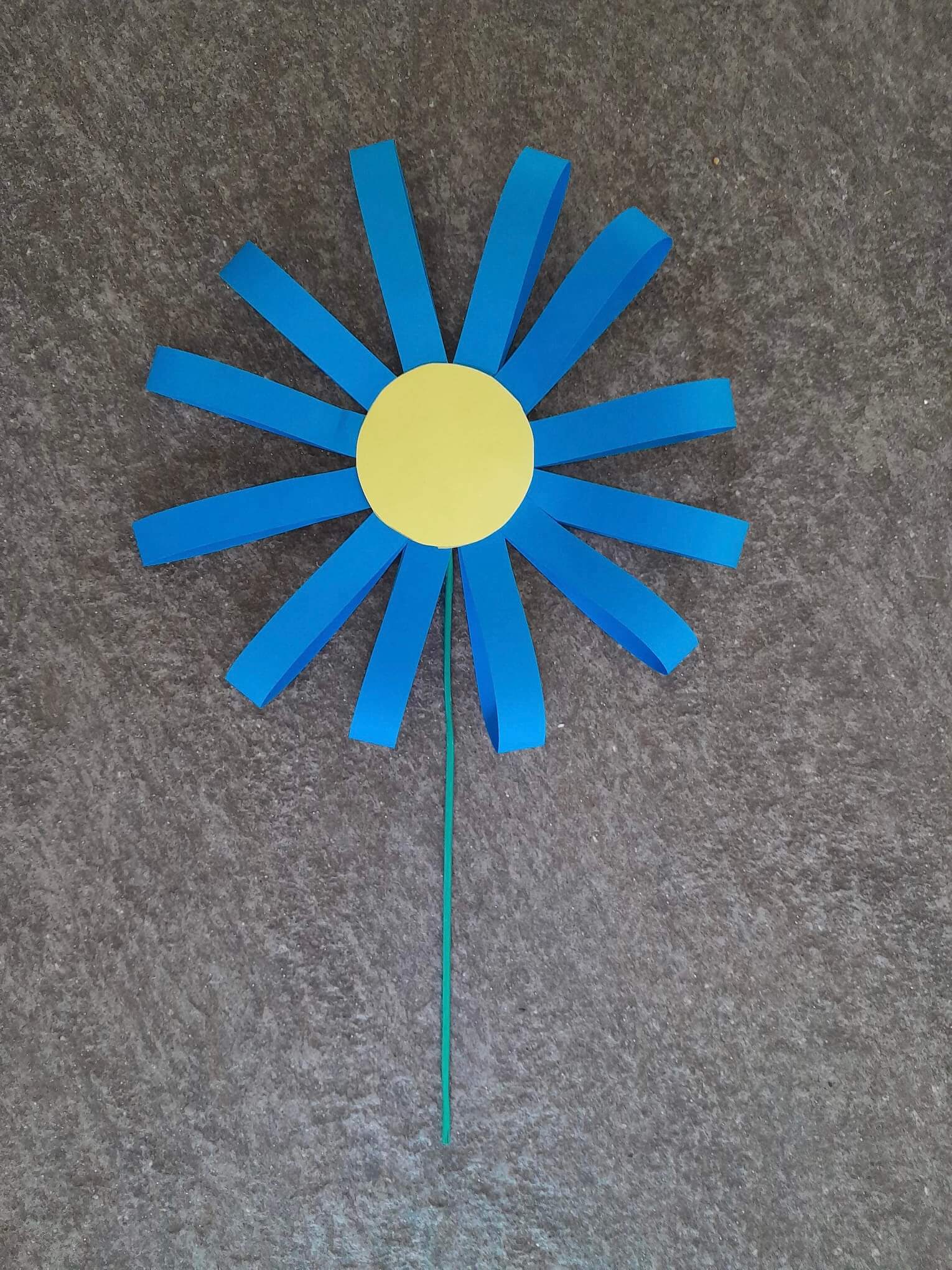 A paper flower with a yellow center, blue petals, and a green stem on a dark textured background. - Home Instead