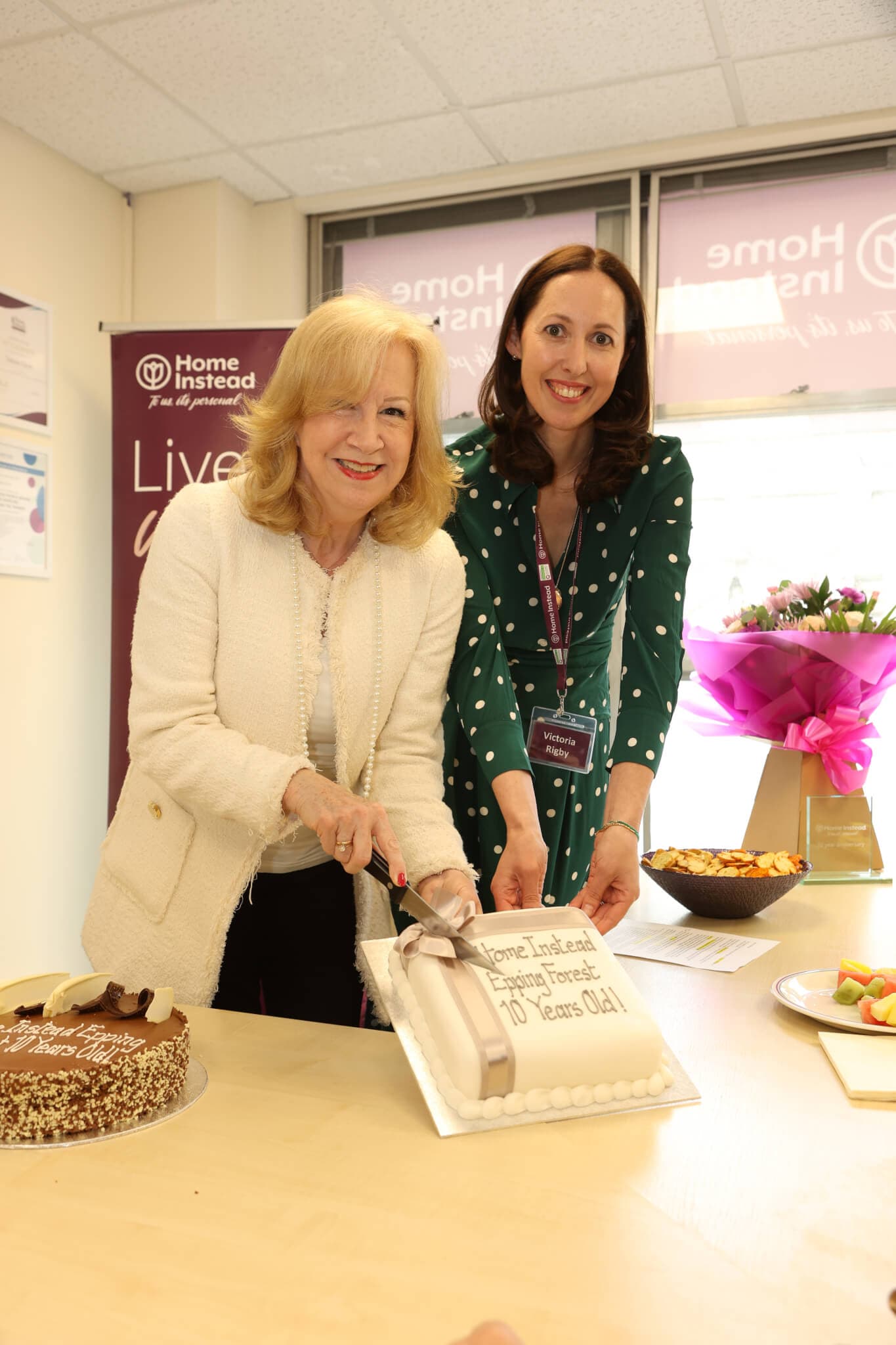 Two women smile while cutting a cake that says "Home Instead Enfield Celebrates 10 Years Old!" in an office setting. - Home Instead
