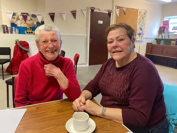 Two older women smiling and seated at a table in a cozy room. A cup of tea is on the table in front of them. - Home Instead