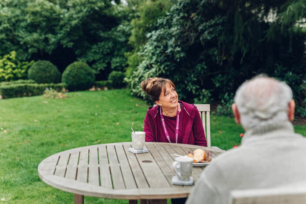 A woman and an older man sit at a wooden table in a garden, smiling and enjoying a conversation and some tea. - Home Instead