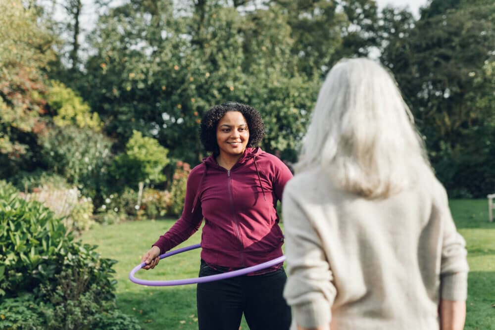 Person in a purple hoodie holds a hula hoop, smiling at another person with gray hair in an outdoor garden setting. - Home Instead