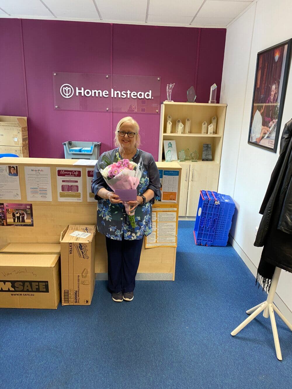 A woman with glasses holding a bouquet of flowers stands smiling in an office space with a "Home Instead" sign. - Home Instead