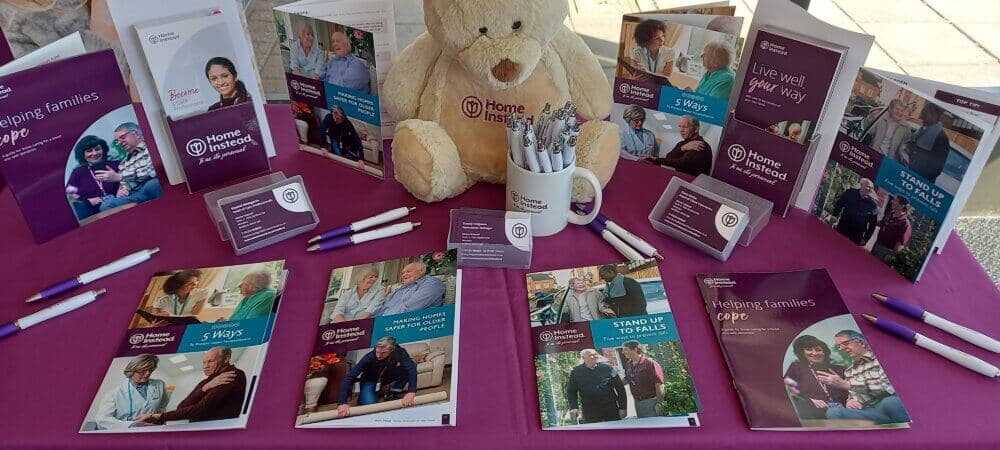 A display table with pamphlets, a teddy bear, pens, and other materials promoting Home Instead senior care services. - Home Instead