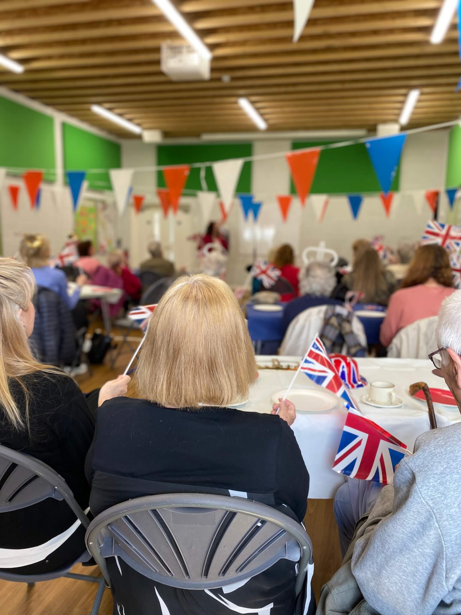 People seated at tables in a festive hall decorated with bunting, holding Union Jack flags and tea on the tables. - Home Instead
