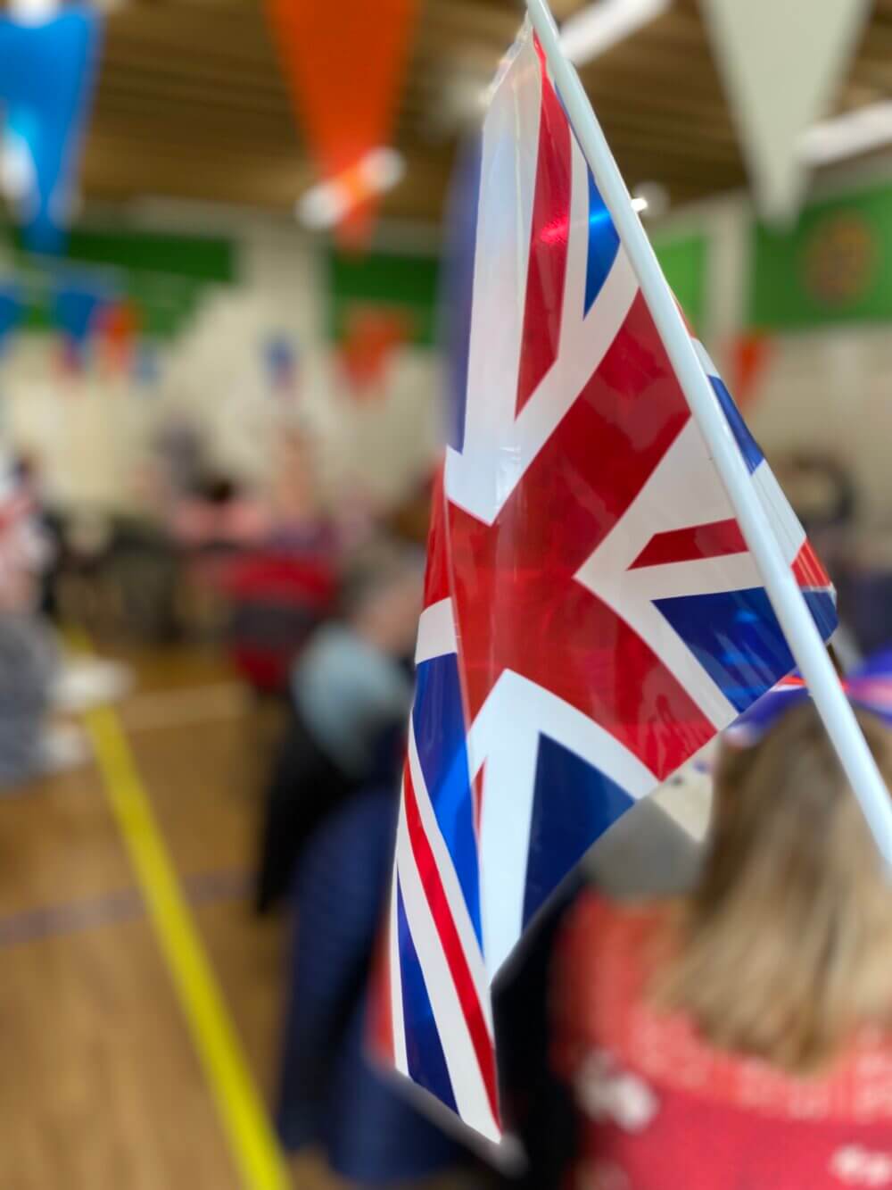 Union Jack flag in focus with blurred background of people and colorful hanging decorations in a hall. - Home Instead