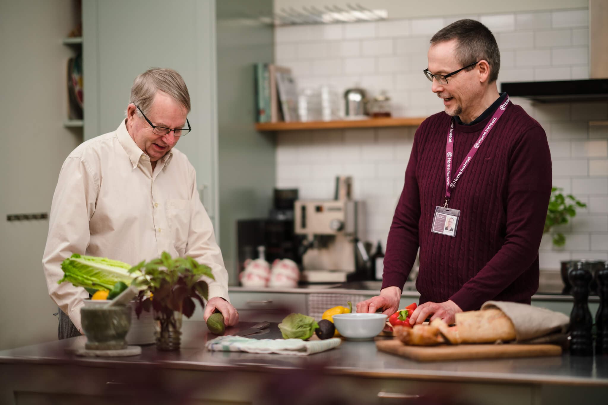 Two men are preparing food together in a modern kitchen, surrounded by fresh vegetables and bread on the counter. - Home Instead