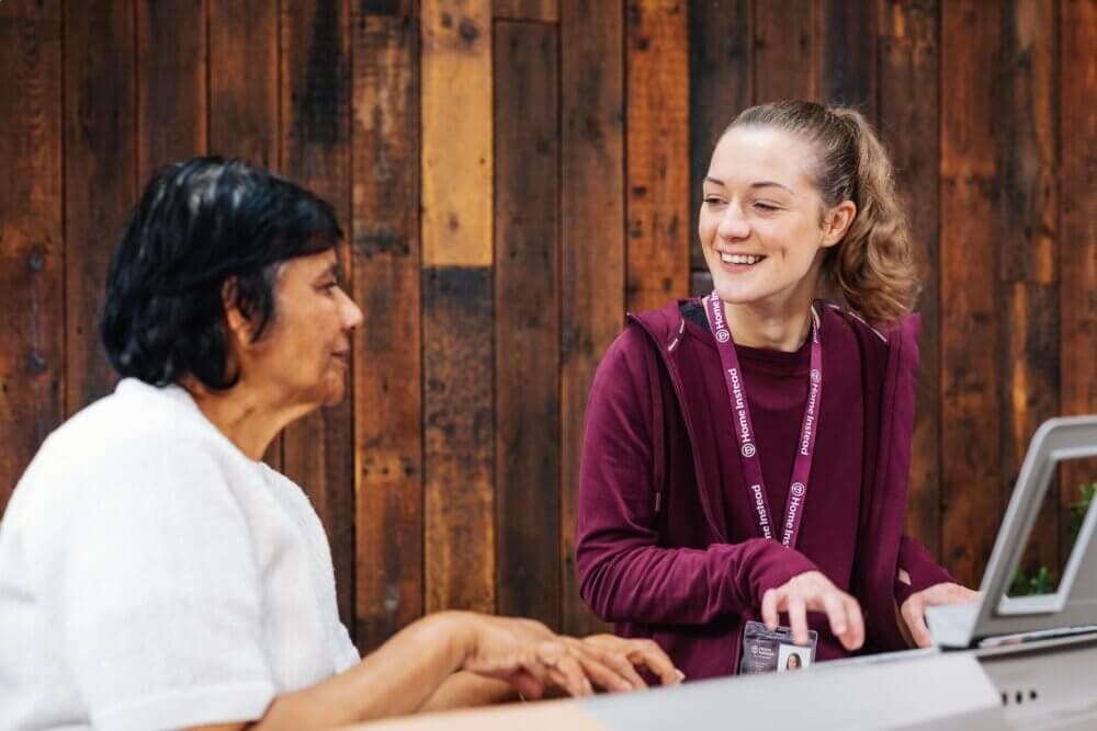 A smiling woman helps another woman with something on a computer. Both are sitting against a wooden wall background. - Home Instead Southampton