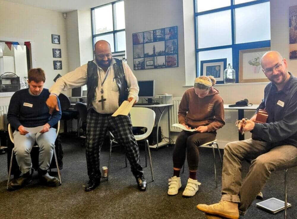 Four people sitting in a room, two reading papers, one playing guitar, and one standing up with an expressive gesture. - Home Instead