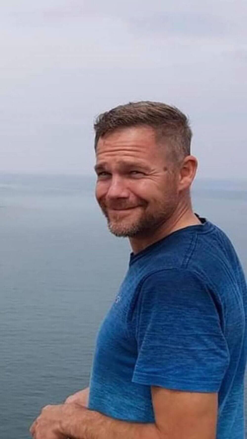 A man with short hair and a beard, wearing a blue shirt, smiling with a body of water in the background. - Home Instead