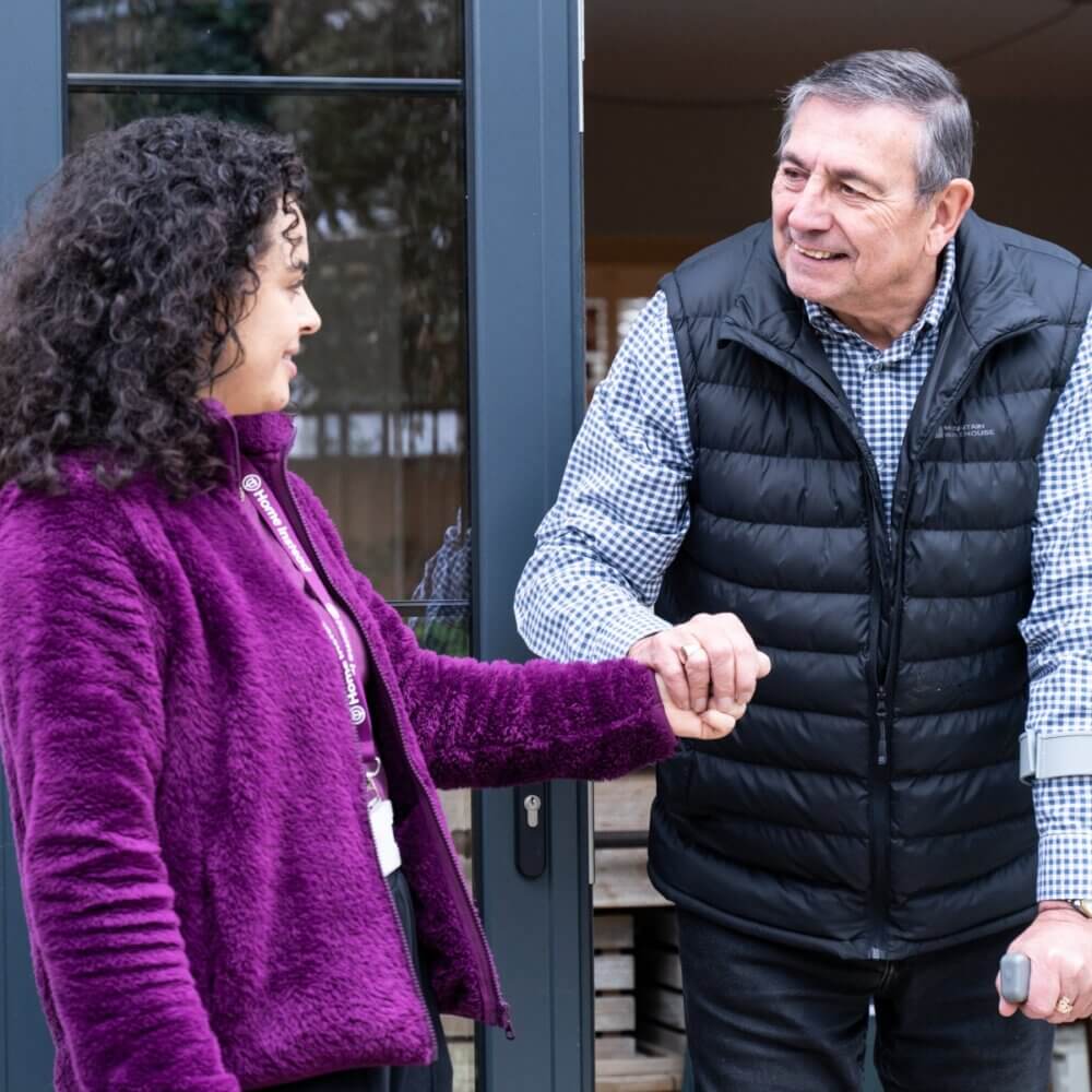 A woman in a purple jacket helps an elderly man with crutches at a doorway, both smiling. - Home Instead
