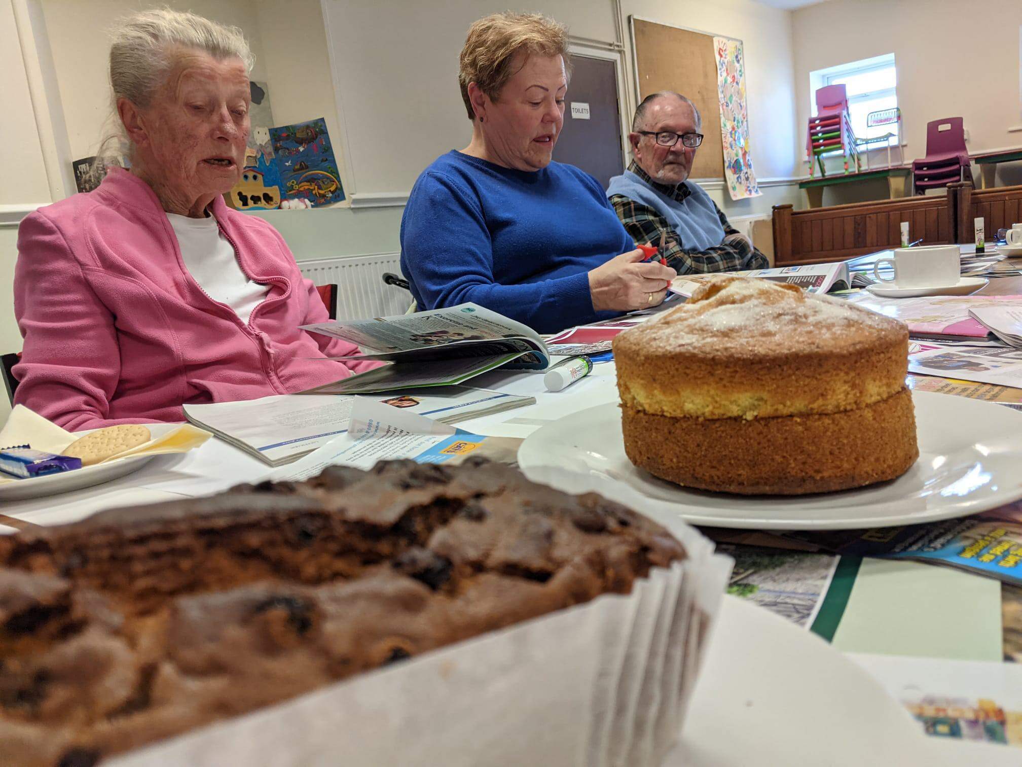 Three people sitting at a table with papers, pens, and cakes, engaging in a discussion or activity in a cozy room. - Home Instead