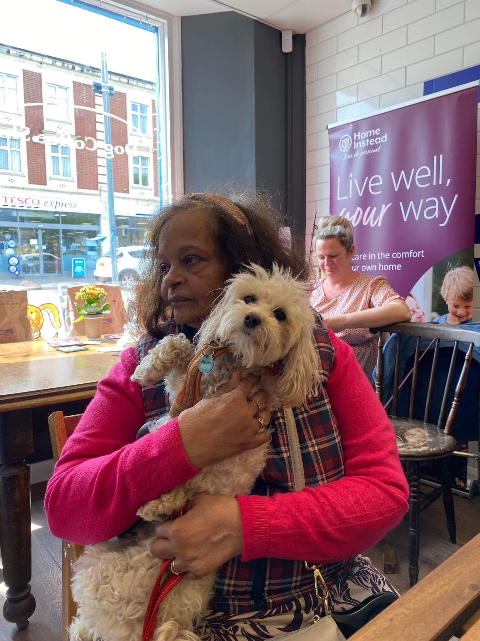 A woman in a bright pink sweater holds a small, fluffy white dog in a cozy cafe setting, with people in the background. - Home Instead
