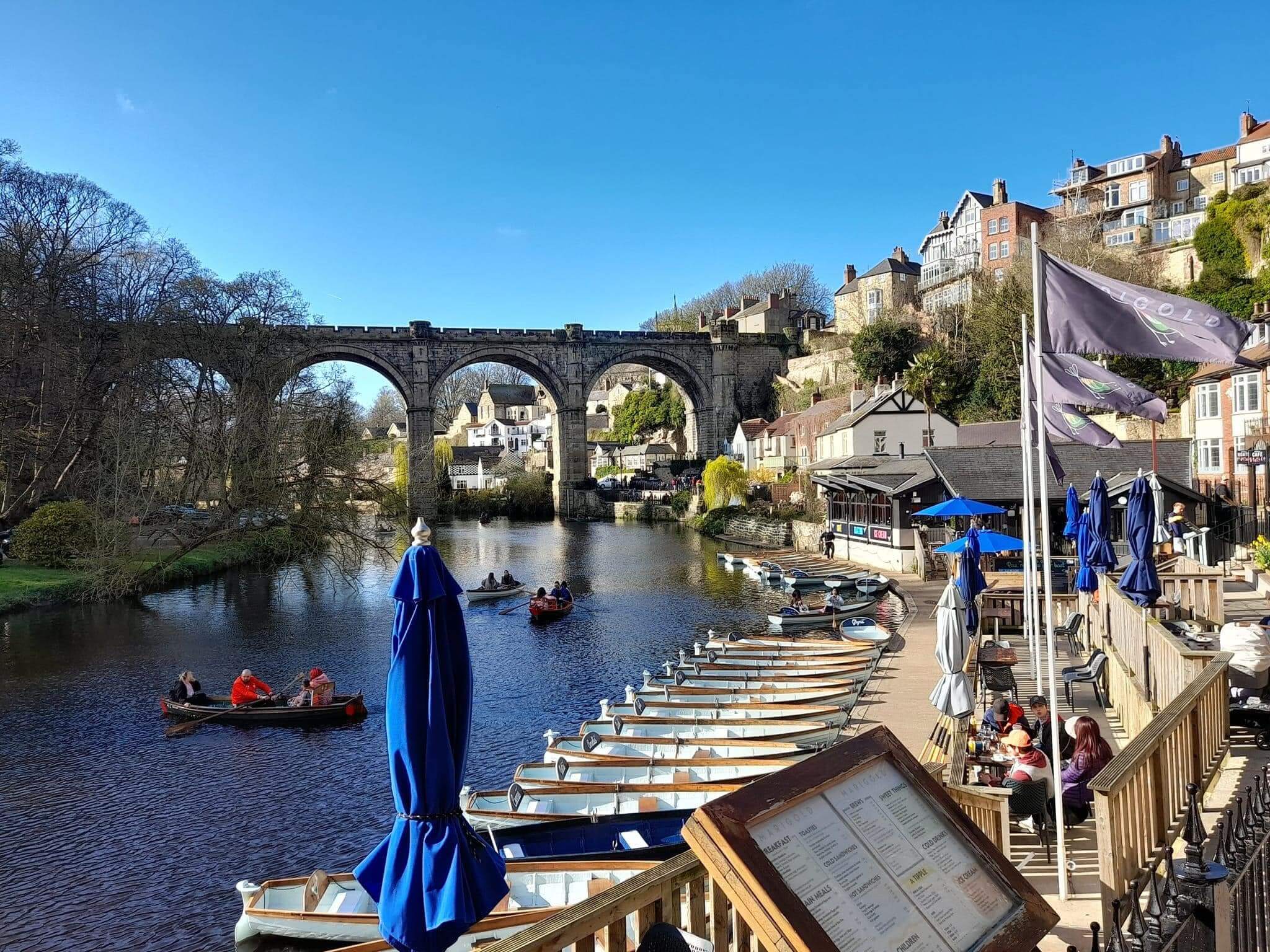 Scenic view of boats on a river, a stone bridge, buildings, and people dining outdoors on a sunny day. - Home Instead