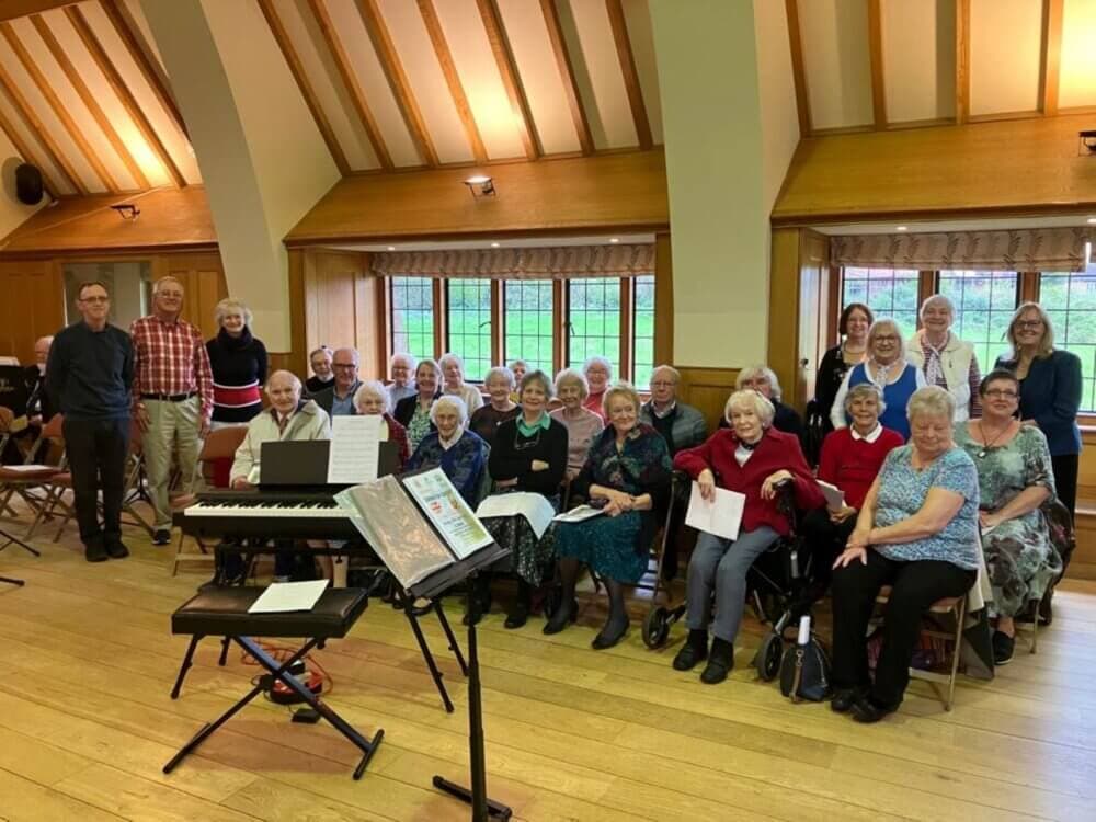 A group of elderly people and a few younger adults smiling and posing in a community room with musical instruments. - Home Instead